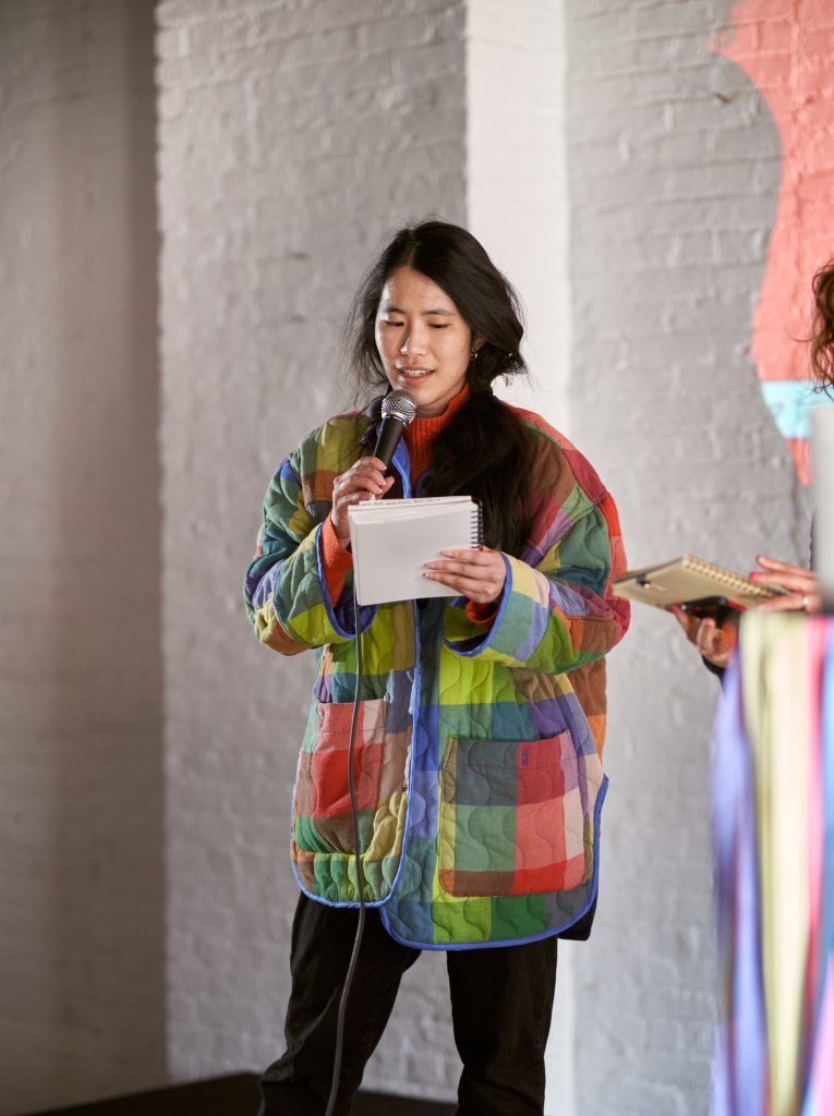 Image of Annie in a multi-colored jacket presenting on stage. She is holding a microphone and looking at a notebook.