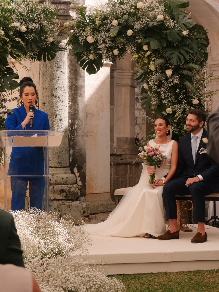 Photo of Annie officiating a wedding. She's wearing a blue suit and has her hair up. The bride and groom sit to her right. Photo by Jenna Ruddock.